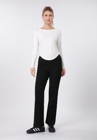 Λευκό τοπ με μακριά μανίκια και στρογγυλεμένη άκρη, συνδυασμένο με μαύρο παντελόνι και μαύρα αθλητικά παπούτσια με λευκές ρίγες. Ομαλές υφές, στενή γραμμή.