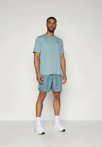 Uomo in piedi rivolto in avanti, indossa una maglietta sportiva azzurro chiaro e pantaloni corti, calze bianche e sneakers bianche, su uno sfondo neutro.