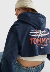 Sk shortená námornícka mikina s okrúhlou kapucňou a grafickým potlačou s nápisom "TOMMY'S Sport & Social Club." Bavlnený materiál, kombinovaný s vysokými džínsami.