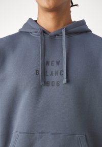 Felpa grigia realizzata in tessuto misto cotone, con tasca a marsupio, lacci e il testo "NEW BALANCE 1906" in nero sul petto.
