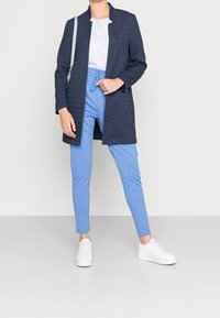 Abrigo largo azul marino con tela texturizada, combinado con pantalones ajustados azul claro y zapatillas blancas. Camiseta simple debajo, diseño minimalista.