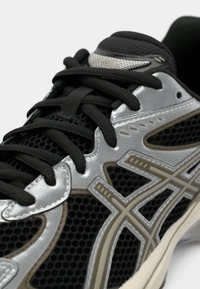 Svart løpesko med mesh-overdel, sølvdetaljer og beige detaljer. Har flate svarte lisser og godt mønstret gummisåle.