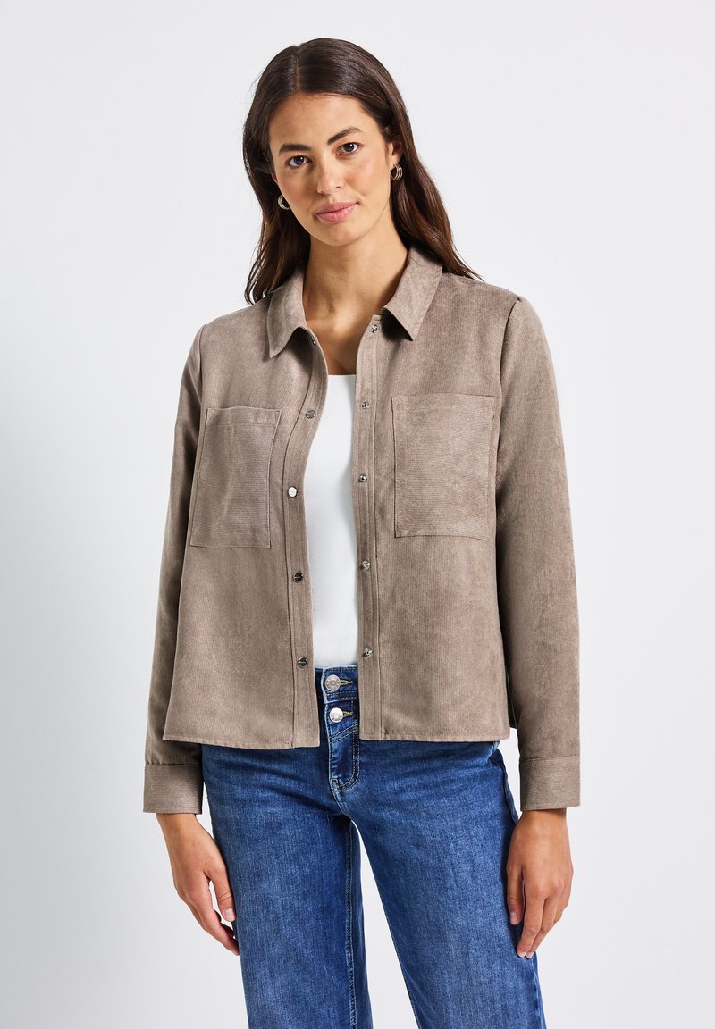 Frau mit beigem Kord-Jackett über einem weißen Top und blauen Jeans, stehend vor einem schlichten weißen Hintergrund.