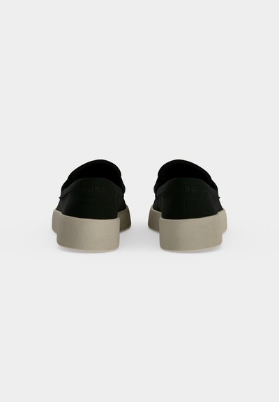 Paire de chaussures noires sans lacets avec semelles beiges épaisses vues de dos sur un fond gris clair uni.