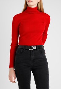 Rode geribbelde coltrui met lange mouwen, gecombineerd met zwarte high-waisted jeans en een zwarte riem met een zilveren ronde gesp.