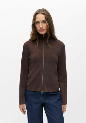 Femme aux longs cheveux bruns portant un pull à col roulé marron zippé et un jean bleu, debout devant un fond blanc uni.