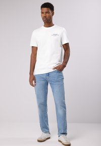 Bílá tričko s krátkým rukávem a grafickým textem, v kombinaci se světle modrými džíny a bílými teniskami s hnědými akcenty. Jednoduchý design, volný střih.