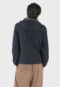 Námořnická modrá lehká bunda s kapucí, s hladkým povrchem a minimalistickým designem. Zadní pohled ukazuje ploché švy a žádná viditelná loga.