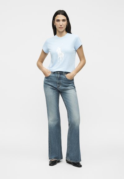 Γυναίκα που στέκεται με τα χέρια στις τσέπες, φοράει ανοιχτό μπλε μπλουζάκι με λευκό λογότυπο, ψηλόμεσο ξεβαμμένο μπλε τζιν και μαύρα παπούτσια.