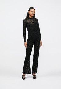 Salopetă din dantelă neagră cu mâneci lungi, model floral transparent, bust ajustat, pantaloni evazați și slituri laterale, asortată cu pantofi negri cu vârf ascuțit.