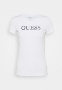 Camiseta blanca de manga corta hecha de algodón con cuello redondo, que presenta un logo brillante en gris "GUESS" impreso en la parte frontal.