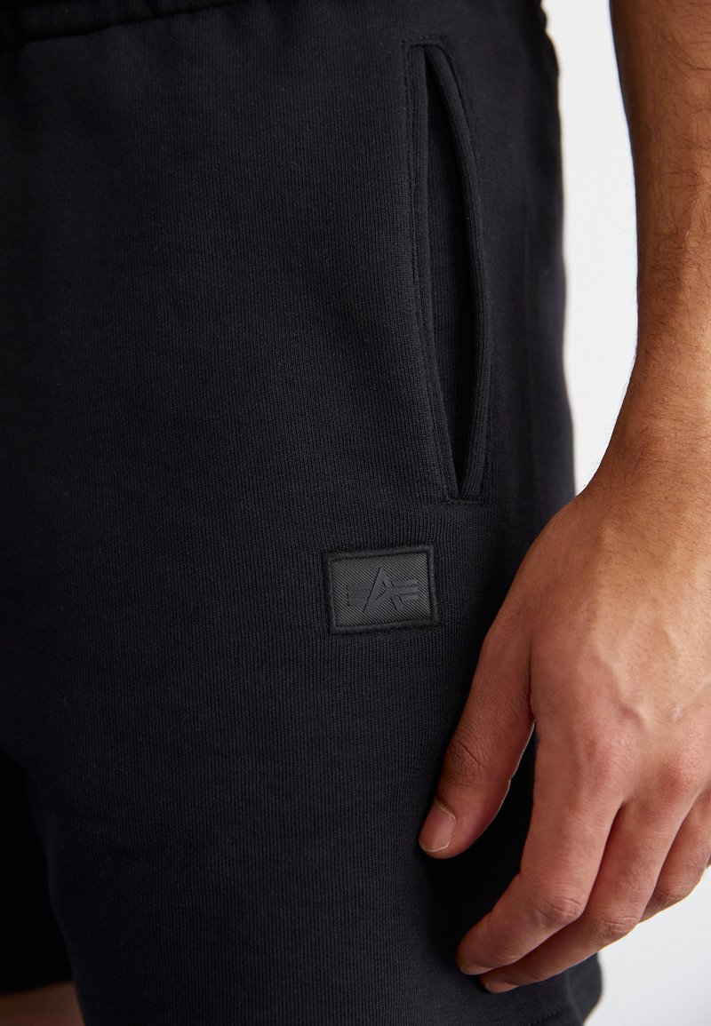 Schwarze Baumwollshorts mit elastischem Bund, ausgestattet mit einer Seitentasche und einem strukturierten schwarzen Logopatch auf dem Stoff.