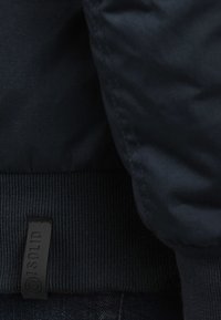 Marineblauwe pufferjack met geribbeld hemband. Close-up toont een gladde stof, subtiele glans en een label met het "J.SOLID" logo.