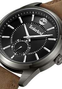 Orologio analogico nero con quadrante testurizzato nero, indicatori argentati, un piccolo sottodial e cinturino in pelle marrone. Presenta un logo Timberland.