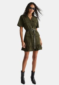 Abito camicia verde oliva con maniche corte, apertura con bottoni, due tasche sul petto, vita con cintura e orlo arricciato. Indossato con stivali neri e occhiali da sole.