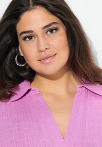 Rosa blus med krage, i en strukturvävd tyg. Modellen har långt brunt hår och silverfärgade hoopörhängen, vänd mot kameran.