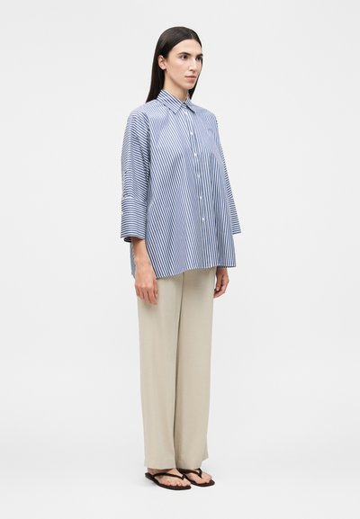 Γυναίκα που στέκεται φορώντας μια ανοιχτή μπλε και άσπρη λευκή ριγέ πουκάμισο με κουμπιά, μπεζ παντελόνια με φαρδιά πόδια και μαύρα σαγιονάρες σε απλό φόντο.