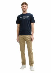 Navyblaues Baumwoll-T-Shirt mit weißem Textlogo, kombiniert mit hellbeigen Chinos und weißen Sneakers. Das Outfit hat einen lässigen Schnitt und kurze Ärmel.