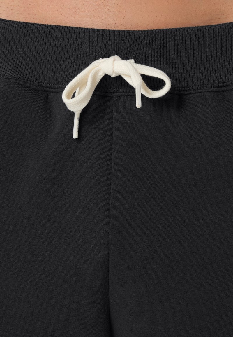 Un pantalon de survêtement noir avec une ceinture côtelée et un cordon blanc noué en nœud. Le tissu semble lisse avec une texture douce.