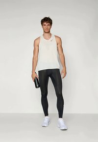 Tânăr îmbrăcat cu un tricou sport alb fără mâneci, colanți de compresie negri, adidași albi, ținând o sticlă de apă neagră, stând pe un fundal gri simplu.