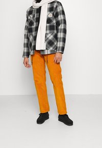 Schwarze und weiße karierte Flanelljacke, orangefarbene Cordhosen und ein leichter Schal, kombiniert mit schwarzen Schuhen. Minimalistischer Hintergrund.