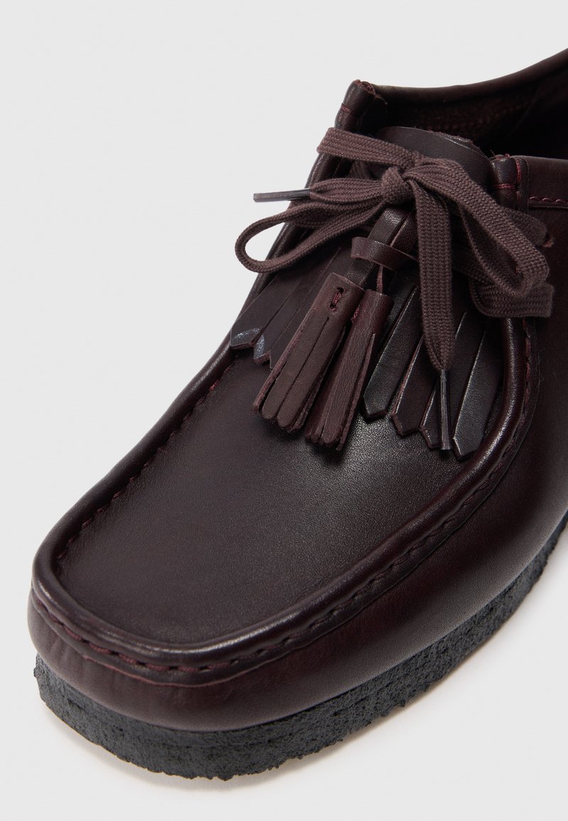 Burgundy leather shoe with raised stitching, decorative tassels, and a textured black rubber sole. Rounded shape with lace closure.