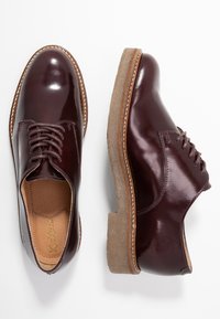 Burgunderfarbene Leder-Schnürschuhe mit glatter Oberfläche, runder Spitze und hellbrauner Gummisohle, die Kontrastnähte zeigt.