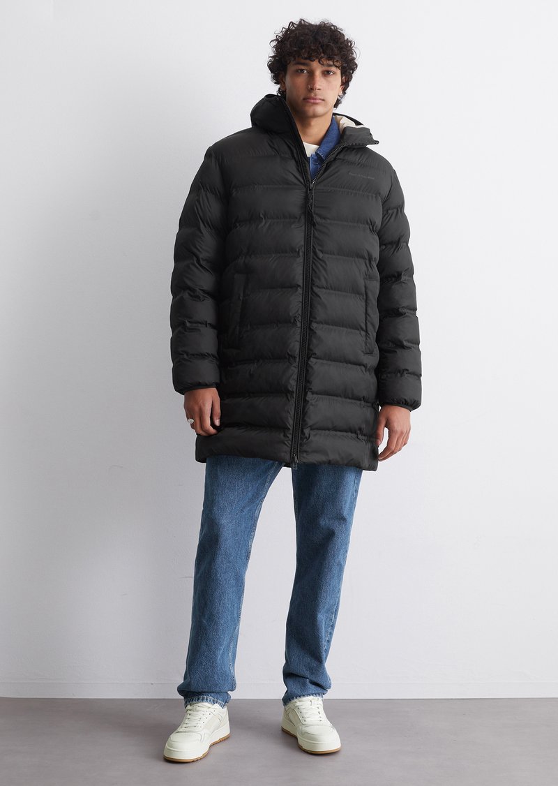 Schwarze Daunenjacke, gestepptes Design, Reißverschluss, hoher Kragen, getragen über einem blauen Hemd und hellblauen Jeans, kombiniert mit weißen Sneakers.