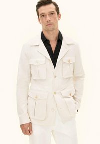 Uomo che indossa una giacca safari color crema con quattro tasche frontali con bottoni sopra una camicia nera, in piedi con una mano in tasca su uno sfondo semplice.