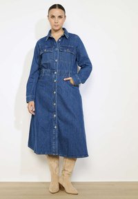 Tumši zila džinsa kleita ar garām piedurknēm, pogu priekšpusi, divām krūšu kabatām un sašaurinātu jostu, apvienota ar līdz ceļiem augstām brūnām zābakiem.