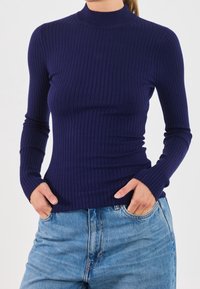 Kvinna i en figurnära marinblå ribbad långärmad tröja och ljusblå högmidjade jeans med händerna i fickorna.