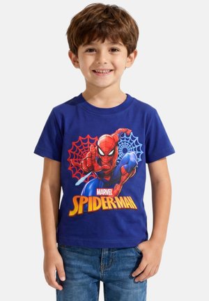 Jeune garçon souriant, portant un t-shirt bleu avec un motif coloré de Spider-Man et un jean bleu, debout sur un fond uni.
