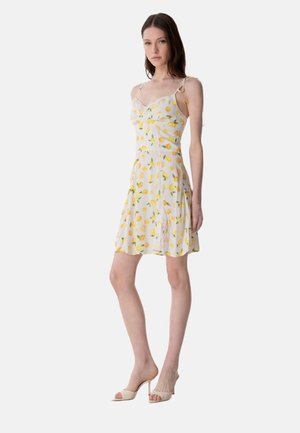 Jeune femme debout, portant une robe blanche sans manches avec des motifs de citrons jaunes et des sandales à talons ouvertes beige.