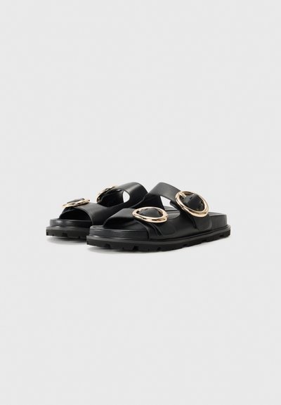 Sorte sandaler med brede krydsremme, gyldne cirkulære metaldele, tekstureret sål og flad design. Fremstillet af syntetisk materiale.