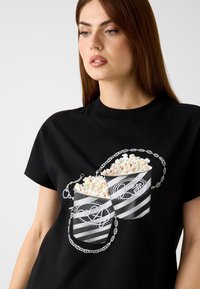 T-shirt nera in cotone con grafica di due contenitori di popcorn a righe con dettagli a catena. Il design include dettagli di popcorn di colore chiaro.