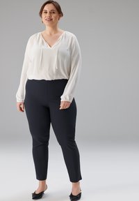 Weiße Bluse mit langen Ärmeln, V-Ausschnitt und Kordelzug am Kragen, kombiniert mit eng anliegenden schwarzen Hosen, die eine glatte Textur und ein klassisches Design aufweisen.