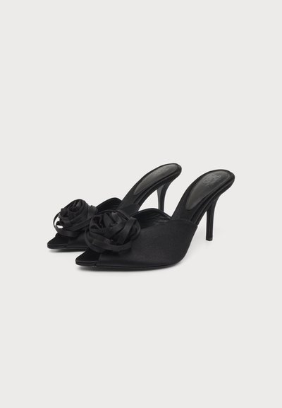 Mules noires à talons hauts et bout ouvert avec un détail de ruban floral à l’avant, présentées sur un fond clair uni.