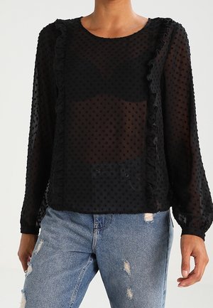 Schwarze, transparente Bluse mit langen Ärmeln, gepunkteter Textur, Rüschensaum und rundem Ausschnitt, kombiniert mit zerrissenen Denim-Jeans.