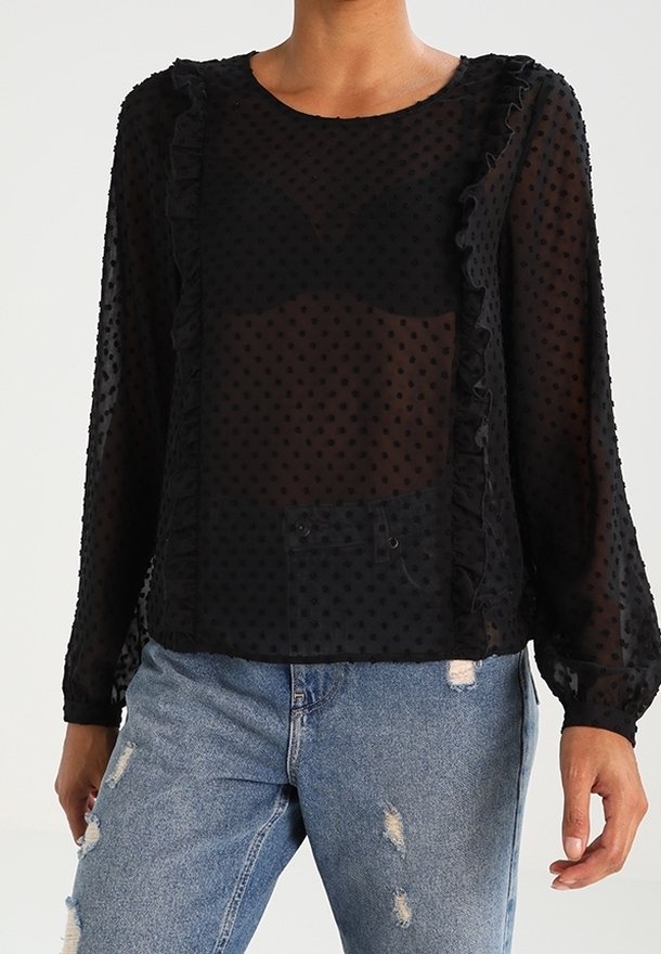 Sort gennemskinnelig bluse med lange ærmer, prikket tekstur, bølgede kanter og rund udskæring, parret med slidte denimjeans.
