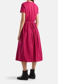 Nainen, jolla on ylleen puettu fuksia midi-mekko, jossa on kerätty vyötärö ja lyhyet hihat, yhdistettynä mustiin nilkkabootseihin, näkyy takaa.