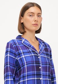 Chemise à carreaux bleus avec un devant à boutons, passepoil blanc au col et une texture douce. Présente différentes nuances de bleu et de violet.