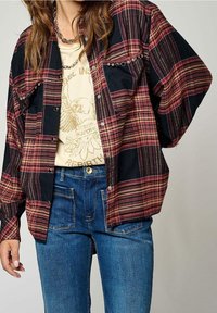 Camicia di flanella a scacchi rossi e blu con bottoni a scatto e dettagli metallici, indossata sopra una t-shirt grafica crema e abbinata a jeans blu.