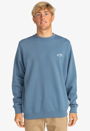 Blue crewneck sweatshirt in soft fabric, featuring long sleeves, ribbed cuffs and hem, with a small white logo on the chest.