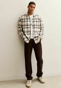 Joven de pie, vistiendo una camisa de cuadros beige sobre una camiseta blanca, pantalones oscuros marrones y zapatillas beige, con las manos en los bolsillos contra una pared lisa.