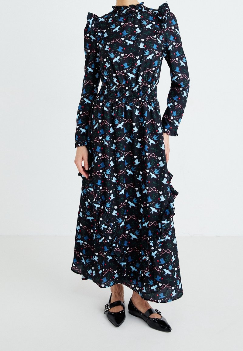 Femme portant une robe midi noire à manches longues avec des imprimés d'oiseaux bleus et de cœurs, des épaules volantées, une taille smockée et des ballerines noires pointues.