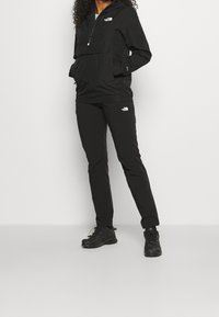 Czarna wiatrówka z półzamkiem, przednią kieszenią i detalem z logo, zestawiona z czarnymi spodniami i czarnymi sportowymi butami. Gładki, lekki materiał.