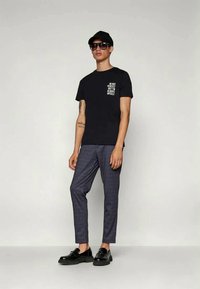 Black t-shirt with white text, gray plaid trousers, black shoes, and sunglasses. Model stands against a neutral background, wearing a black cap.