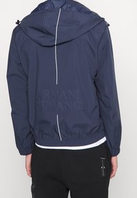 Námořnická bunda s kapucí, která má na zadní straně bílou svislou linku, elastický pas a embosované logo "ARMANI EXCHANGE". Hladká textura.
