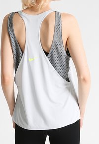 Lichtgrijs sporttanktop met grijze mesh achterkant, voorzien van een reflecterende gele Nike-logo. Mouwloos ontwerp en losse pasvorm voor comfort.