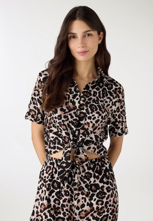 Giovane donna con lunghi capelli scuri che indossa una camicia a maniche corte con stampa leopardata annodata e pantaloncini abbinati, in piedi davanti a uno sfondo bianco.